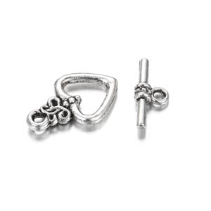 May include: A silver-toned heart-shaped toggle clasp with an ornate design and a matching bar component. The clasp features a detailed floral-like embellishment on the heart. The bar has a loop for attachment. Ideal for jewelry making.