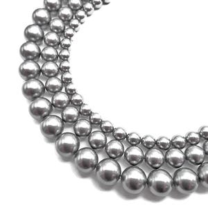 May include: Close-up of several strands of gray pearl necklaces. The pearls vary in size, creating a layered effect. The pearls have a lustrous, reflective surface, and are set against a white background.
