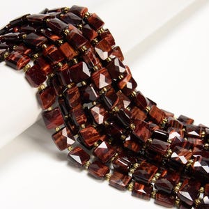 May include: Close-up of a strand of faceted, rectangular beads in shades of deep red and brown. The beads are strung together with small gold-colored spacers, creating a layered effect. The gemstones have a polished, reflective surface.