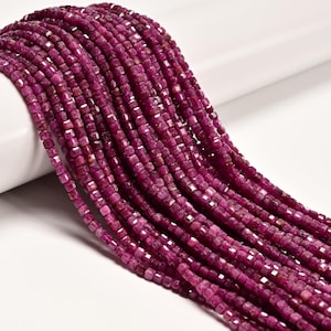 May include: A collection of multiple strands of small, faceted, deep magenta-colored square beads. The beads are arranged in parallel lines, showcasing a uniform shape and a textured, reflective surface across the entire length of the strands.