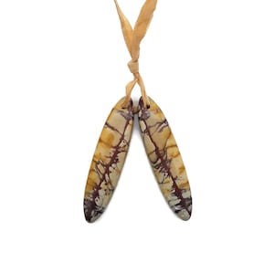 May include: Two elongated, oval-shaped pendants with a beige and brown marbled pattern, connected by a tan ribbon. The pendants have a polished finish and a natural stone appearance, ideal for jewelry making or crafting.