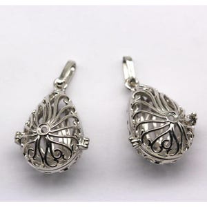 May include: Two silver-colored, teardrop-shaped pendants. Each pendant features an open filigree design with a small loop at the top for a chain. The pendants have a hinged closure and a spherical interior. The background is plain white.