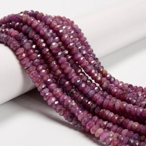 May include: Strands of faceted, deep magenta-colored gemstone beads. The beads are strung together, creating a textured and visually appealing display. The gemstones have a polished, reflective surface, and are arranged against a white background.