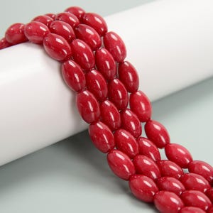 May include: Close-up of strands of oval-shaped, deep red beads. The beads are strung together, creating a flowing, textured pattern. The beads have a glossy finish, reflecting light. The background is a white, curved surface.