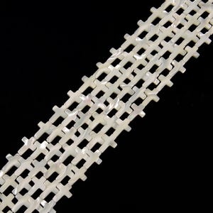 May include: Close-up of several strands of white cross-shaped beads. The beads have a pearlescent sheen and are arranged diagonally against a black background. The crosses are uniform in size and shape, creating a repeating pattern. Ideal for jewelry making.
