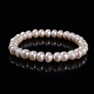 May include: A white pearl bracelet with a stretch design. The pearls are round and have a slight luster. The bracelet is displayed on a black surface, creating a contrast that highlights the jewelry.