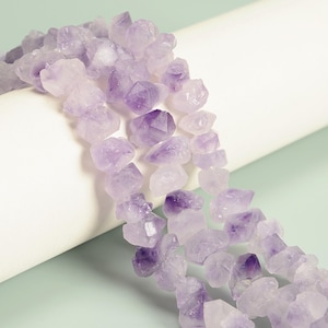 May include: Close-up of strands of raw amethyst crystal beads. The beads are a mix of light and deep purple hues, with a rough, natural texture. The crystals are arranged in a cascading pattern against a white and light green background.