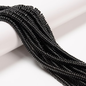 May include: Close-up of multiple strands of glossy black beads. The beads are small, cylindrical, and strung together, creating a textured, layered effect. The beads are displayed against a white background.