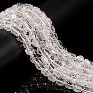 May include: Strands of clear, oval-shaped quartz beads. Each bead is strung on a white cord, creating a layered effect. The beads have a polished, translucent appearance, reflecting light. The background is black, highlighting the beads.