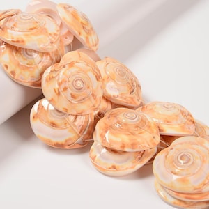 May include: A collection of polished, spiral-shaped seashell beads in shades of cream and brown. The beads are strung together, showcasing their natural patterns and textures. Ideal for jewelry making or coastal-themed crafts.