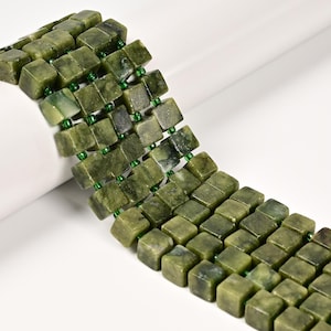 May include: Close-up of a bracelet made of square, olive-green jade beads. The beads are strung together with small, green beads. The bracelet is displayed on a white surface, showcasing the natural variations in the jade.