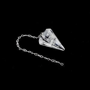 Puede incluir: Un péndulo de cristal transparente con un diseño facetado en forma de cono. Una delicada cadena plateada está unida al cristal. El péndulo está sobre un fondo negro.