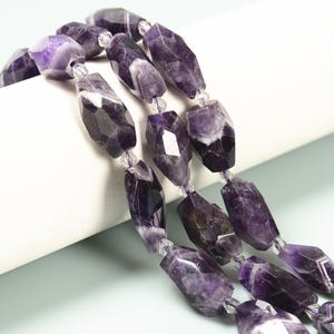 May include: Strands of faceted amethyst gemstone beads. The beads are a deep purple color with white inclusions and are strung on a clear cord. The gemstones have an irregular, elongated shape and are displayed against a white background.