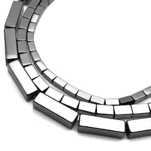 May include: Close-up of three strands of rectangular, metallic gray beads. The beads are uniform in size and shape, with a polished, reflective surface. The beads are strung together, creating a necklace or bracelet.