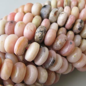 May include: A collection of round, flat, disc-shaped beads in various shades of pale pink, peach, and cream. Many of the beads feature natural inclusions, dark grey or black speckles, and subtle variations in opacity and surface texture.