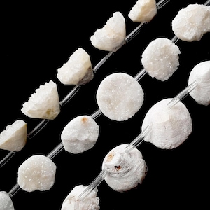 May include: Strands of white, irregular-shaped beads with a crystalline texture. The beads are strung on clear, thin cords against a black background. Some beads are round, others are more angular.
