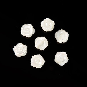 May include: Seven white, rose-shaped beads are arranged against a black background. The beads have a pearlescent sheen, with detailed petal formations. These decorative beads are ideal for jewelry making or craft projects.