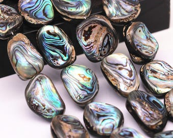Op de afbeelding: Een streng iriserende abalone schelp kralen, elke kraal is een unieke ovale vorm met een glad, gepolijst oppervlak. De kralen zijn een mix van blauwe, groene en gouden kleuren.