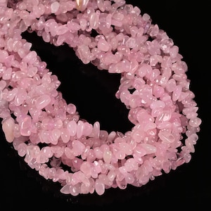May include: Strands of light pink, irregular-shaped rose quartz beads. The beads are strung together, creating a necklace-like arrangement. The background is black, which highlights the color and texture of the gemstones. The beads are likely used for jewelry making.