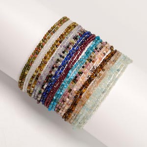 May include: A stack of colorful gemstone bracelets, each made with small, round beads. The bracelets are arranged in a rainbow pattern, with shades of blue, green, pink, brown, and white.