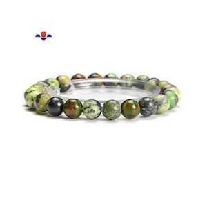 May include: A beaded bracelet featuring a variety of green, brown, and black spherical beads. The beads have a speckled appearance and are strung together on a clear elastic band. The bracelet is displayed on a clear, circular stand.