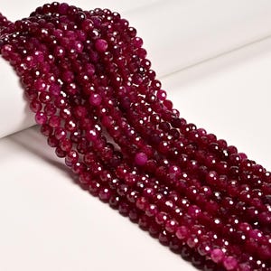 May include: Strands of faceted, deep red gemstone beads. The beads have a polished, reflective surface, and are strung together. The beads are a rich, dark red color, and are arranged in a cascading pattern. The background is white.