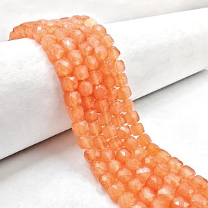 May include: Close-up of strands of faceted, orange-colored beads. The beads are square-shaped with multiple facets, creating a sparkling effect. The beads are strung together, forming a flexible strand. The background is a white, rolled surface.