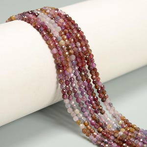 May include: Strands of faceted gemstone beads in shades of pink, purple, and brown. The beads are strung together, creating a flowing, textured appearance. The gemstones have a sparkling, polished finish, reflecting light.