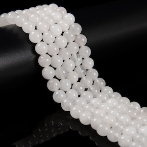 May include: Strands of white, round gemstone beads. The beads are smooth and have a slightly translucent appearance. The beads are strung together, creating a flowing, organic shape. The background is black.