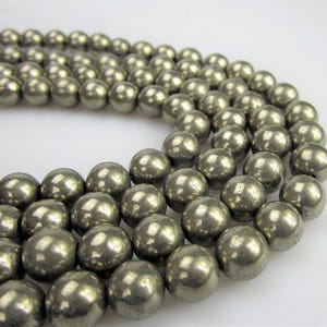 May include: Close-up of multiple strands of round, metallic-looking beads. The beads have a silvery-gold color and a smooth, reflective surface. The beads are arranged in a curved pattern, suggesting they are part of a necklace or bracelet. The background is white.