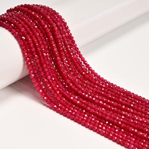 May include: Strands of faceted, deep red beads cascade across a white background. The beads are small and evenly sized, reflecting light with a sparkling effect. The image showcases the beads' rich color and texture, ideal for jewelry making.