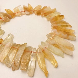 May include: A necklace composed of raw, golden-yellow citrine crystal shards. Each shard is strung together with a thin red thread, creating a circular shape. The crystals vary in size and shape, displaying a natural, unpolished appearance.
