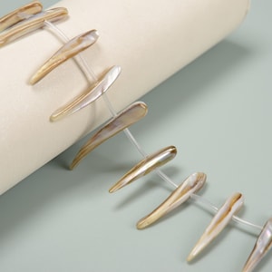 Puede incluir: Primer plano de un collar de cuentas de concha alargadas e iridiscentes. Las cuentas son una mezcla de colores crema, beige y nacarado, ensartadas con hilo transparente. Las cuentas están dispuestas en diagonal sobre un fondo crema.