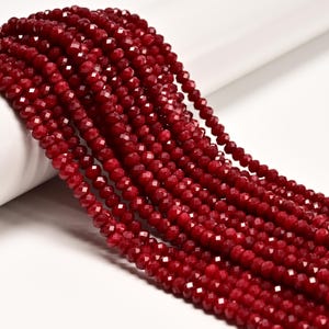 May include: Multiple strands of deep red, faceted crystal beads arranged in parallel lines. Each individual bead features a multi-sided cut that reflects light, creating a sparkling appearance across the entire collection of dark crimson jewelry components.