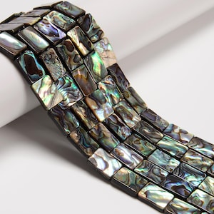 May include: Close-up of rectangular abalone shell beads with iridescent colors of green, blue, and brown. The beads are strung together, showcasing their polished, rectangular shape. Ideal for jewelry making.