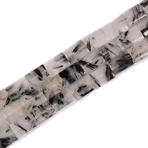 May include: Close-up of a strand of square-shaped beads. The beads are clear with black, needle-like inclusions. The beads are arranged diagonally against a white background, showcasing their unique patterns and textures.