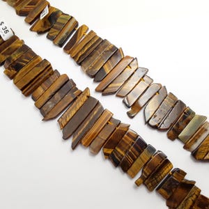 May include: Two strands of tiger eye gemstone beads. The beads are rectangular and have a mix of brown, gold, and black colors. The beads are strung together with a small hole drilled through each one. The background is white.