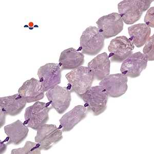 May include: Strands of raw amethyst gemstone beads. The beads are a light purple color and have a rough, natural texture. They are strung on a purple cord. The image is a close-up shot, highlighting the details of the gemstones.