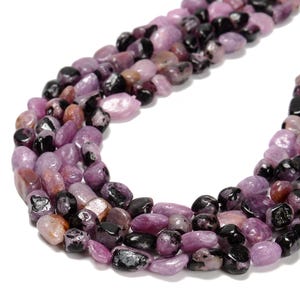 May include: Close-up of strands of tumbled gemstones in shades of purple, black, and brown. The stones are smooth and rounded, with a polished appearance. The gemstones are strung together, creating a necklace or bracelet.