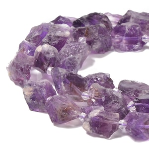 May include: A close-up of a strand of raw amethyst gemstone beads. The beads are a deep purple color with rough, natural edges. The beads are strung together with clear string and small, clear beads.