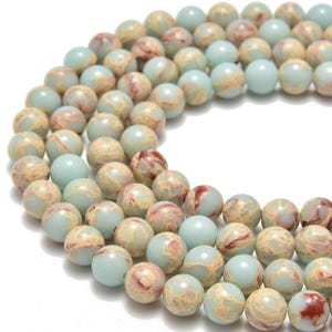 May include: Strands of round gemstone beads in shades of pale blue, tan, and burgundy. The beads have a marbled appearance, with variations in color and pattern. These beads are suitable for jewelry making.