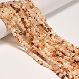 May include: A collection of round, polished beads arranged in multiple parallel strands. The beads feature a smooth, iridescent surface with a color palette ranging from creamy off-white and soft beige to warm shades of light and dark brown.