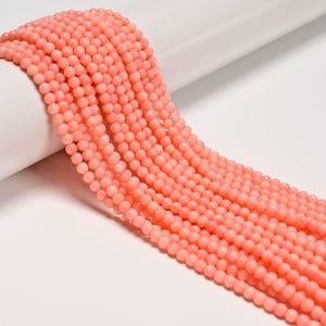 May include: Close-up of strands of small, round, coral-colored beads. The beads are uniform in size and shape, and are strung together. The beads are arranged in a flowing pattern, suggesting they are ready for jewelry making.
