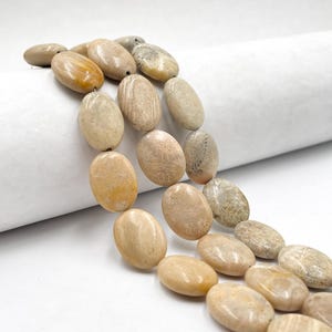 May include: Strand of oval-shaped, beige fossil coral beads. The beads have a polished, smooth surface with natural variations in color and texture. The beads are strung together, ready for jewelry making or crafting projects.