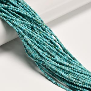 May include: A long strand of small, round turquoise beads featuring a vibrant cyan blue color with natural dark brown and black matrix veining throughout. The beads are uniform in size and arranged in a dense, multi-layered cluster.