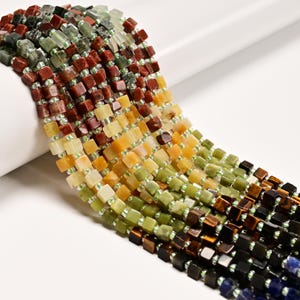 May include: Close-up of strands of colorful gemstone beads. The beads are square-shaped and come in a variety of colors, including green, red, yellow, brown, and blue. The beads are strung together with small, clear beads.