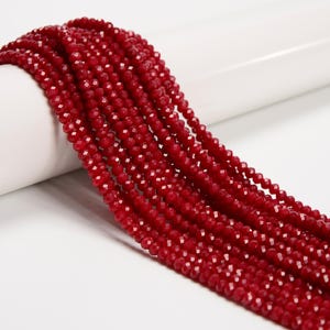 May include: Strands of faceted, deep red beads. The beads are small and uniform in size, with multiple strands cascading down. The beads have a sparkling, reflective quality, suggesting they are made of glass or a similar material. The background is white.