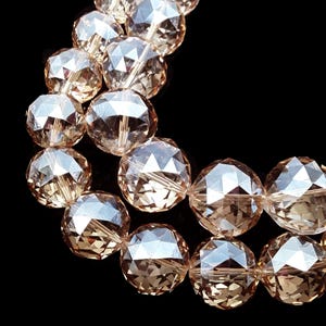 May include: Close-up of faceted, amber-colored beads. The beads are arranged in a curved pattern against a black background. Each bead has multiple reflective surfaces, creating a sparkling effect. The beads are likely for jewelry making.