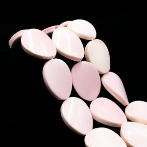 May include: Strand of oval-shaped, light pink beads with a glossy finish. The beads are strung together, creating a curved arrangement against a black background. The beads vary slightly in color, with some appearing more white.