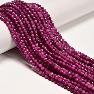 May include: A long strand of small, round, faceted beads in a deep magenta-pink color. The beads are uniform in size and shape, featuring a reflective, multi-faceted surface that catches the light, arranged in a dense, continuous string.